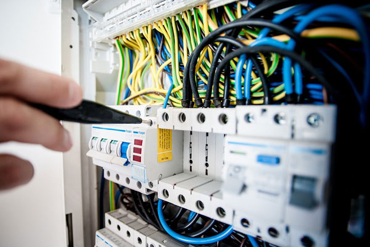 A close-up photograph of an electrician's hand using a screwdriver to work on a complex electrical panel filled with colorful wiring and circuit breakers